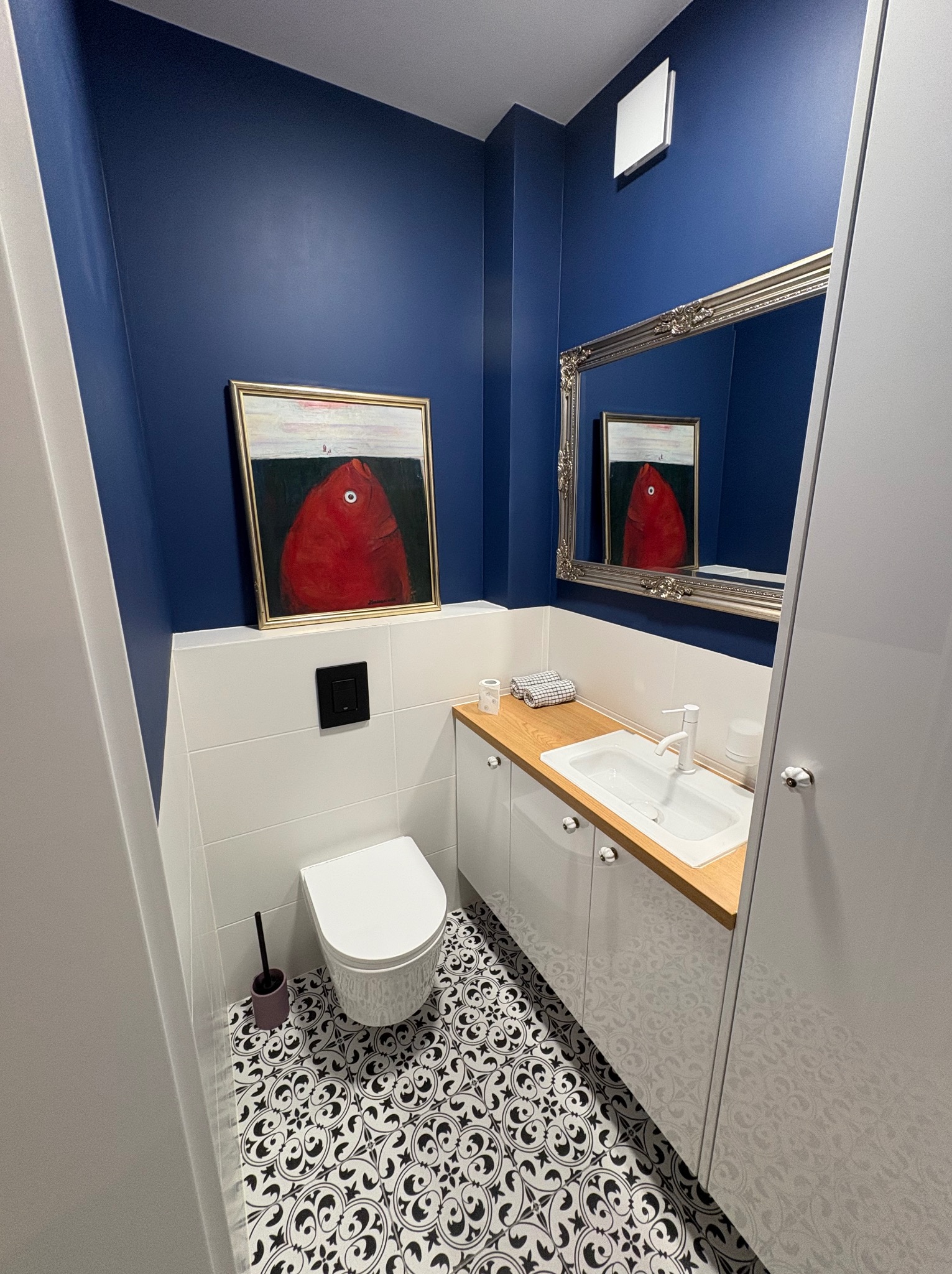 Niebiesko-biała łazienka z wzorzystą podłogą, lustrem w ozdobnej ramie i obrazem czerwonej ryby. Umywalka z drewnianym blatem i białe szafki.