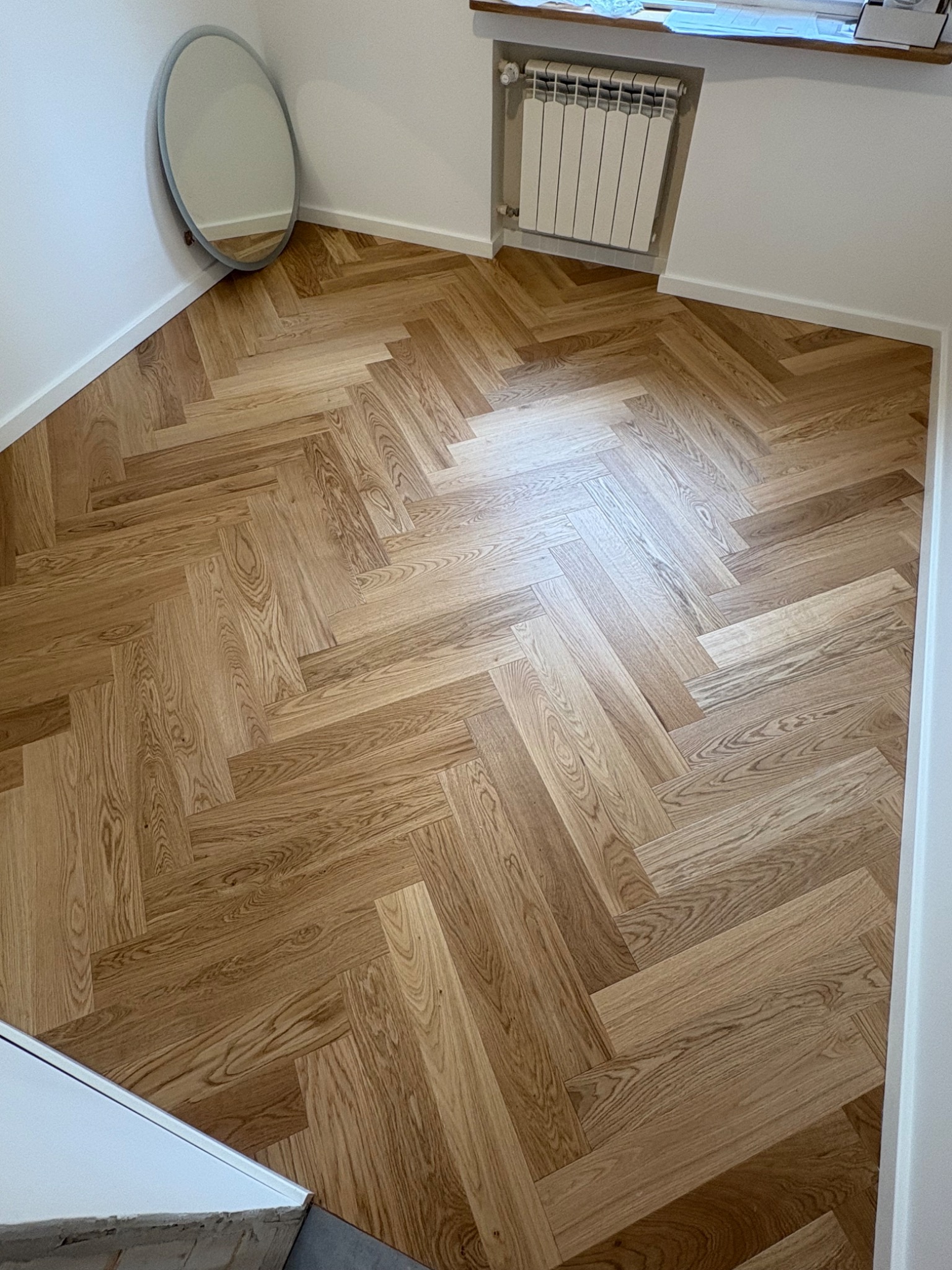 Parkiet ułożony w jodełkę w jasnym wnętrzu, z owalnym lustrem na ścianie i grzejnikiem pod oknem. Widok na podłogę po wykończeniu.