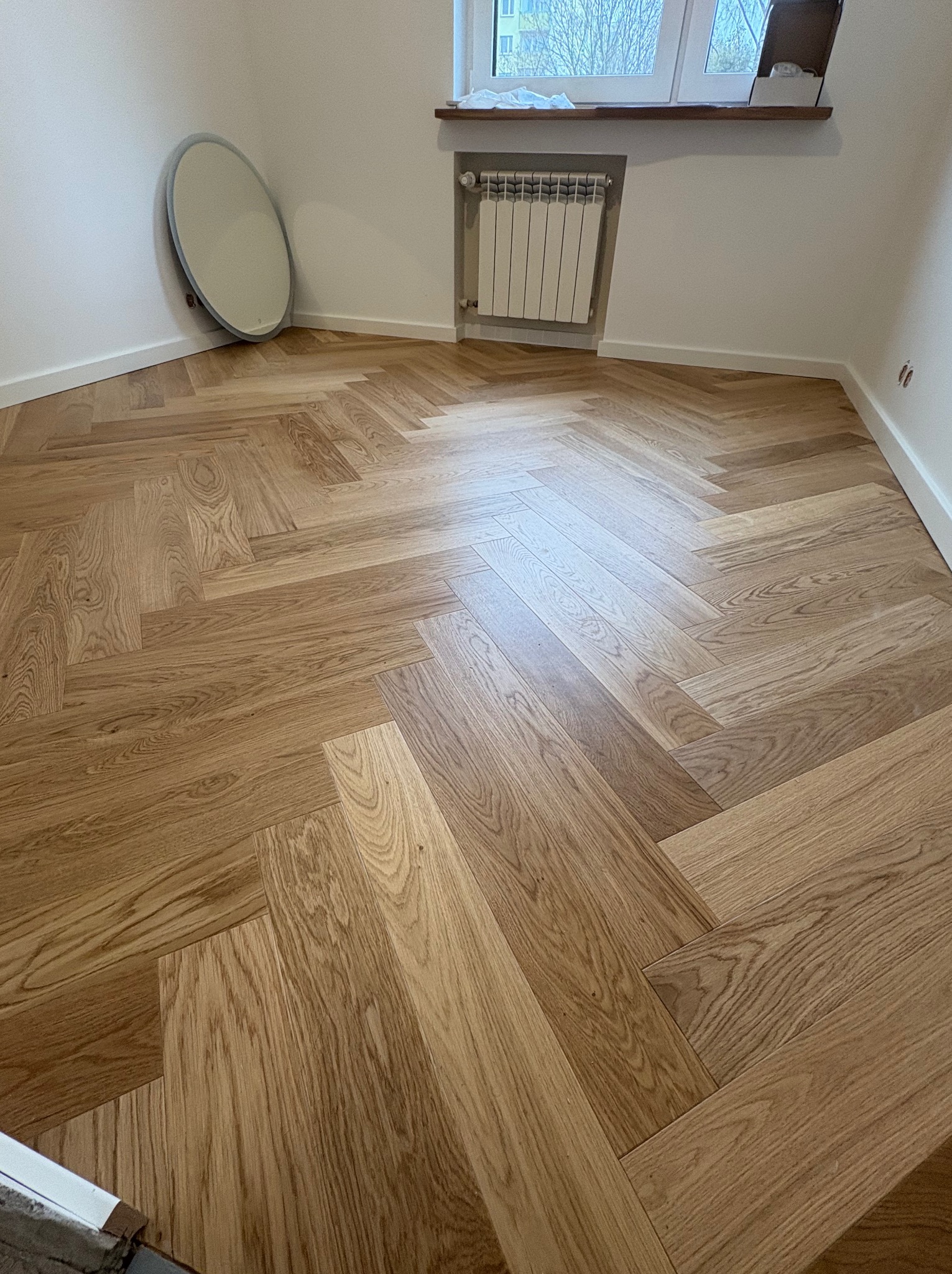 Parkiet ułożony w jodełkę francuską, widok na fragment pokoju z grzejnikiem i lustrem opartym o ścianę. Jasne drewno, naturalne oświetlenie.