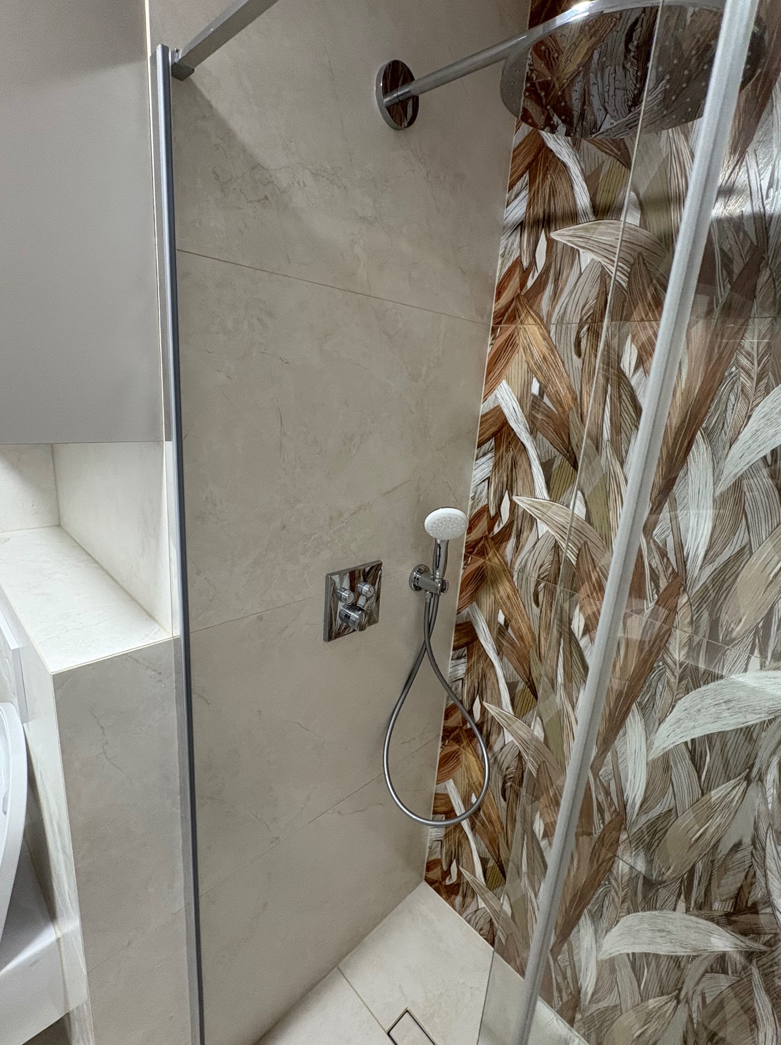 Wykończona kabina prysznicowa z beżowymi płytkami imitującymi marmur i dekoracyjną ścianą z motywem liści. Widoczna bateria i deszczownica.