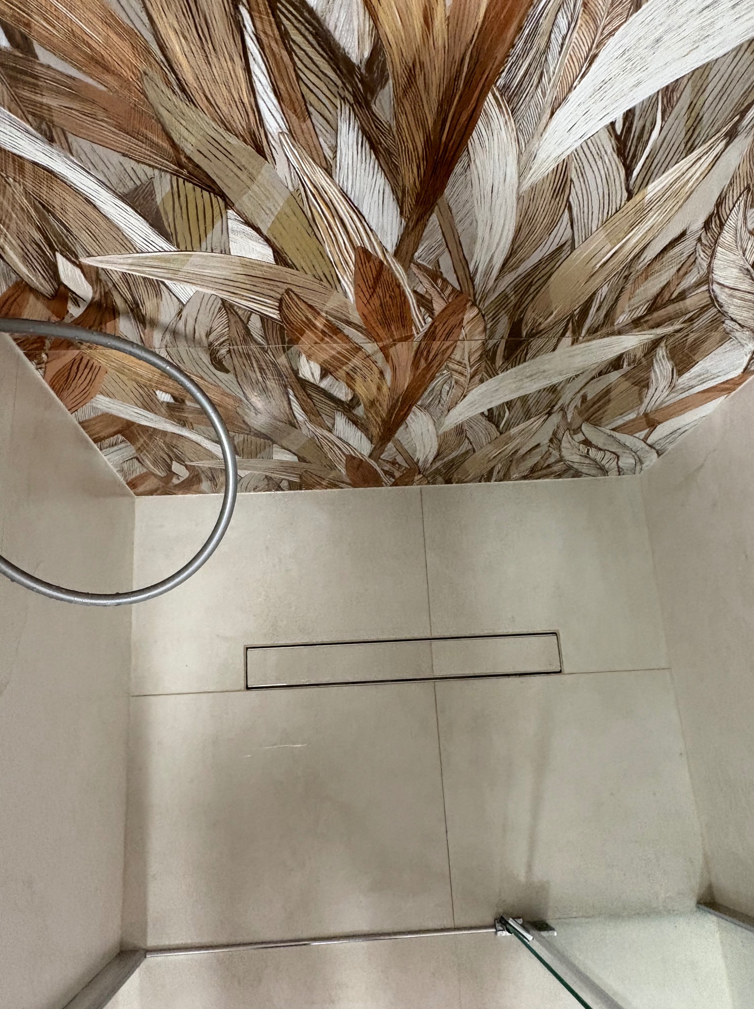 Beżowa łazienka z wzorzystym sufitem w odcieniach brązu i beżu. Widoczny odpływ liniowy, uchwyt na ręcznik i fragment szklanej kabiny. Nietypowe wykończenie sufitu.