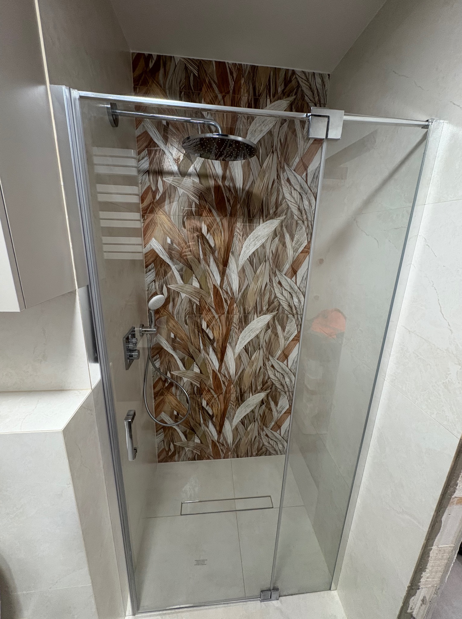 Kabina prysznicowa z deszczownicą i wzorzystą ścianą w odcieniach brązu i beżu. Widoczny odpływ liniowy i chromowana armatura. Wykończona łazienka.