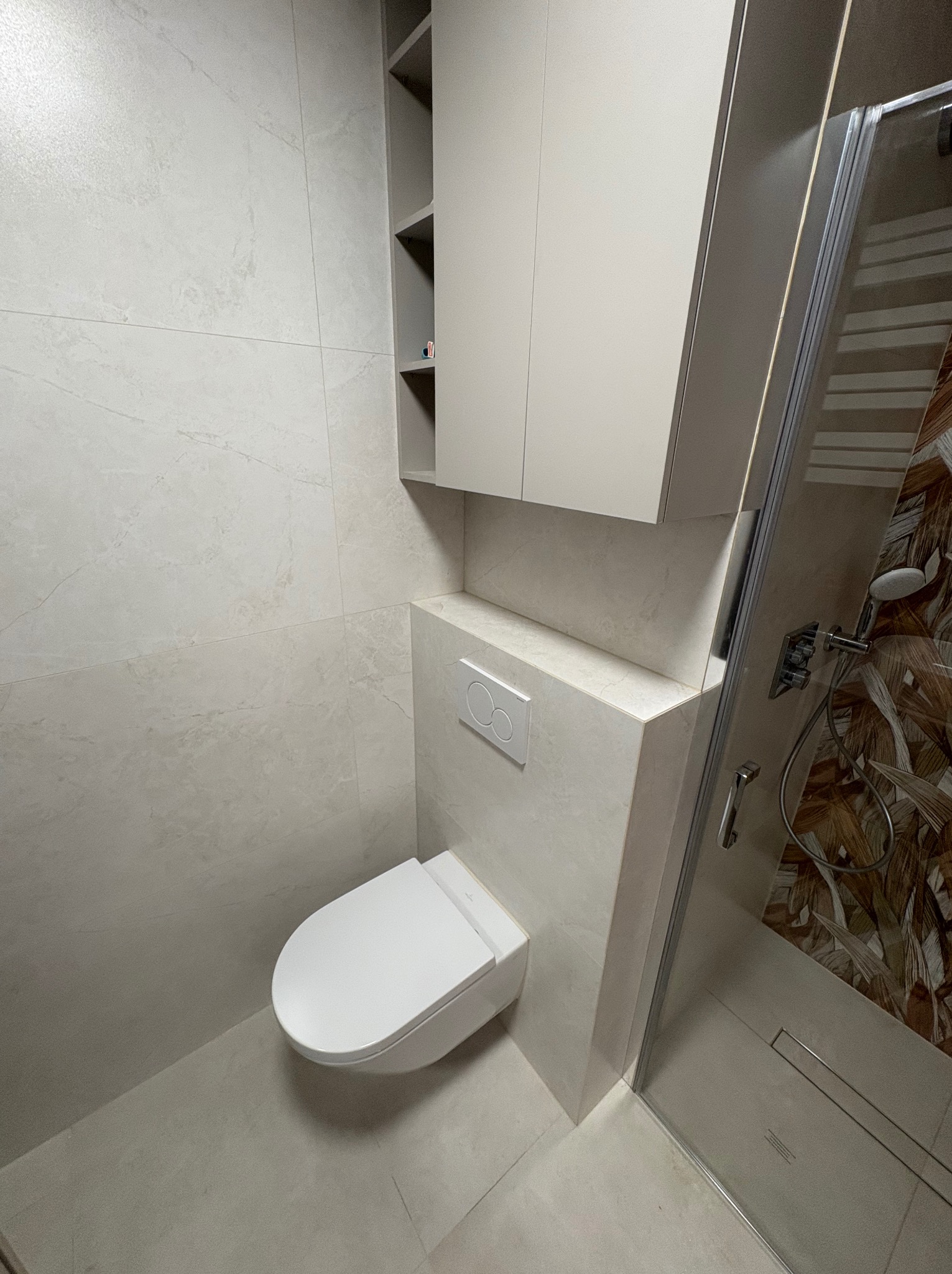 Nowoczesna łazienka z podwieszaną toaletą, szafką nad nią i kabiną prysznicową z wzorzystą ścianą. Jasne płytki na ścianach i podłodze. Minimalistyczny design.