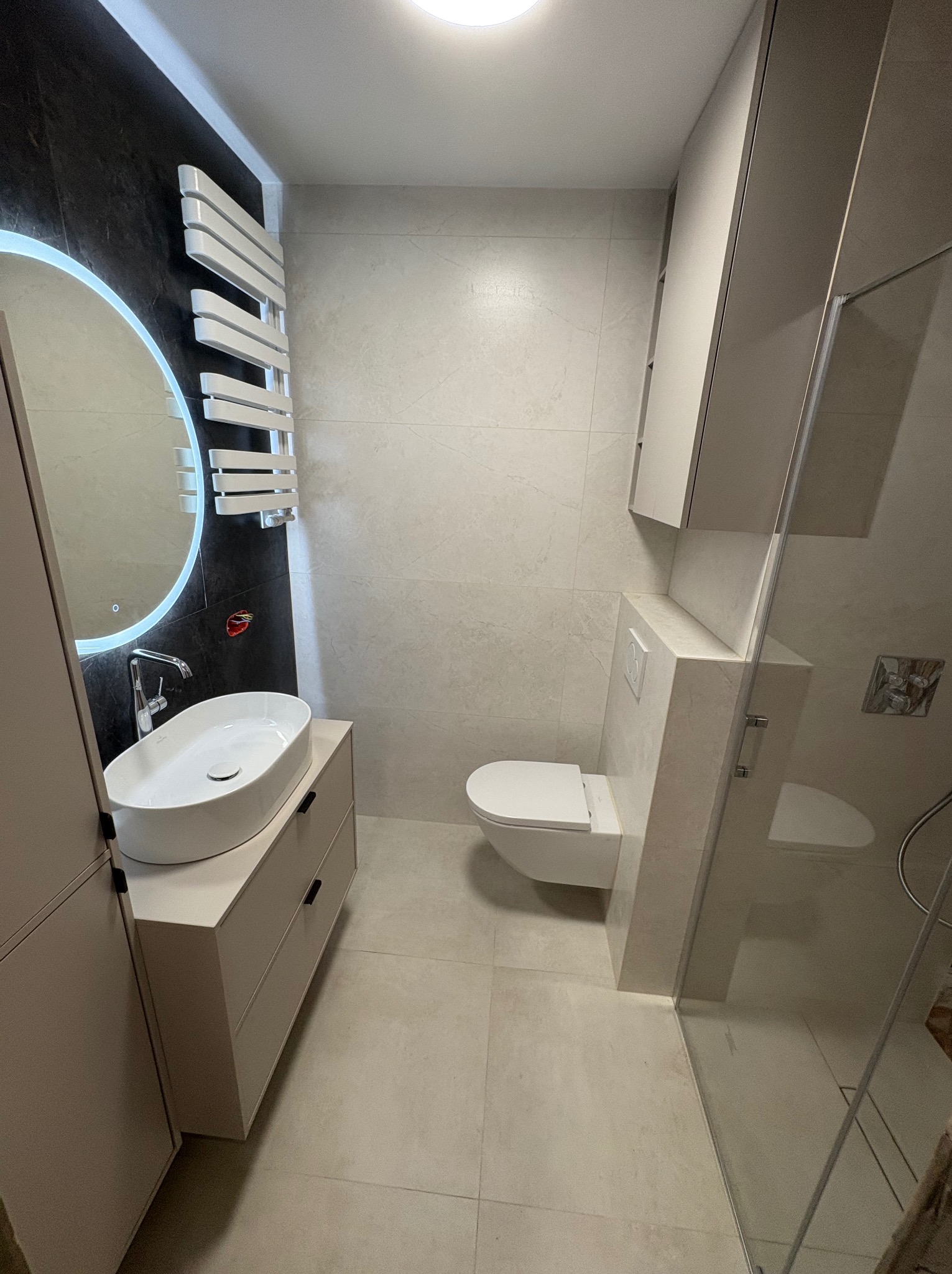 Nowoczesna łazienka z owalną umywalką na szafce, podświetlanym lustrem i grzejnikiem drabinkowym na tle ciemnej ściany. Jasne płytki i kabina prysznicowa.