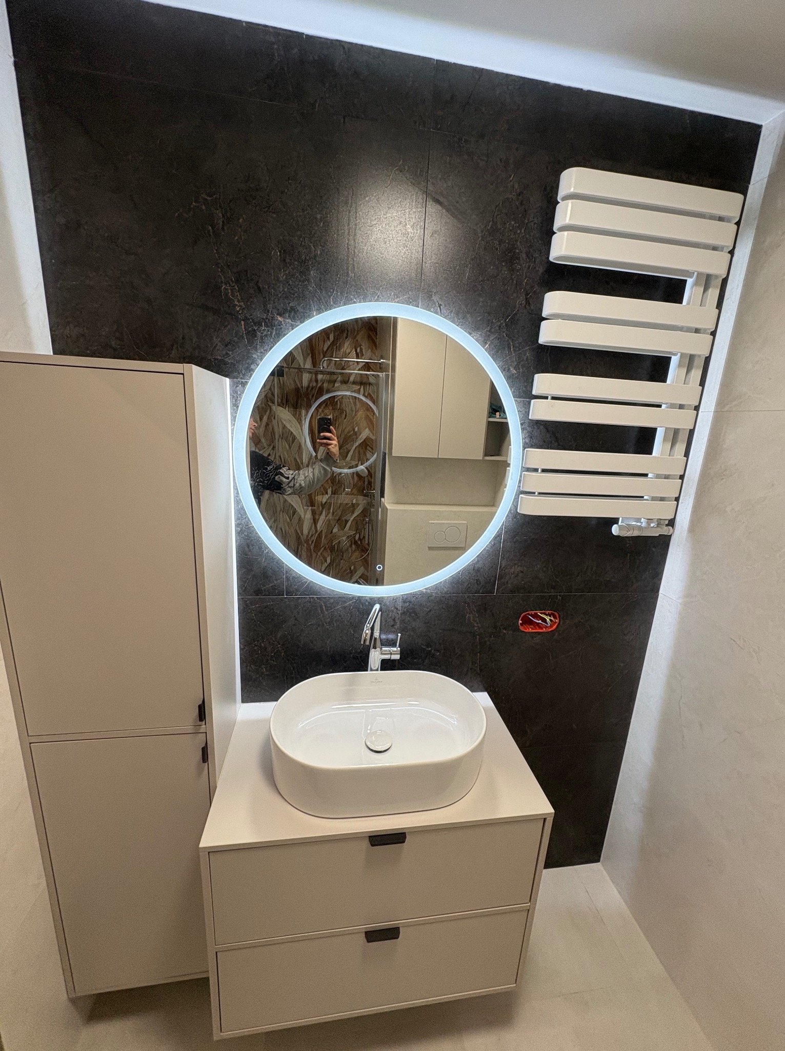 Nowoczesna łazienka z okrągłym lustrem LED, białą umywalką nablatową i grzejnikiem drabinkowym na tle ciemnej, marmurkowej ściany. Minimalistyczny design, jasne meble.