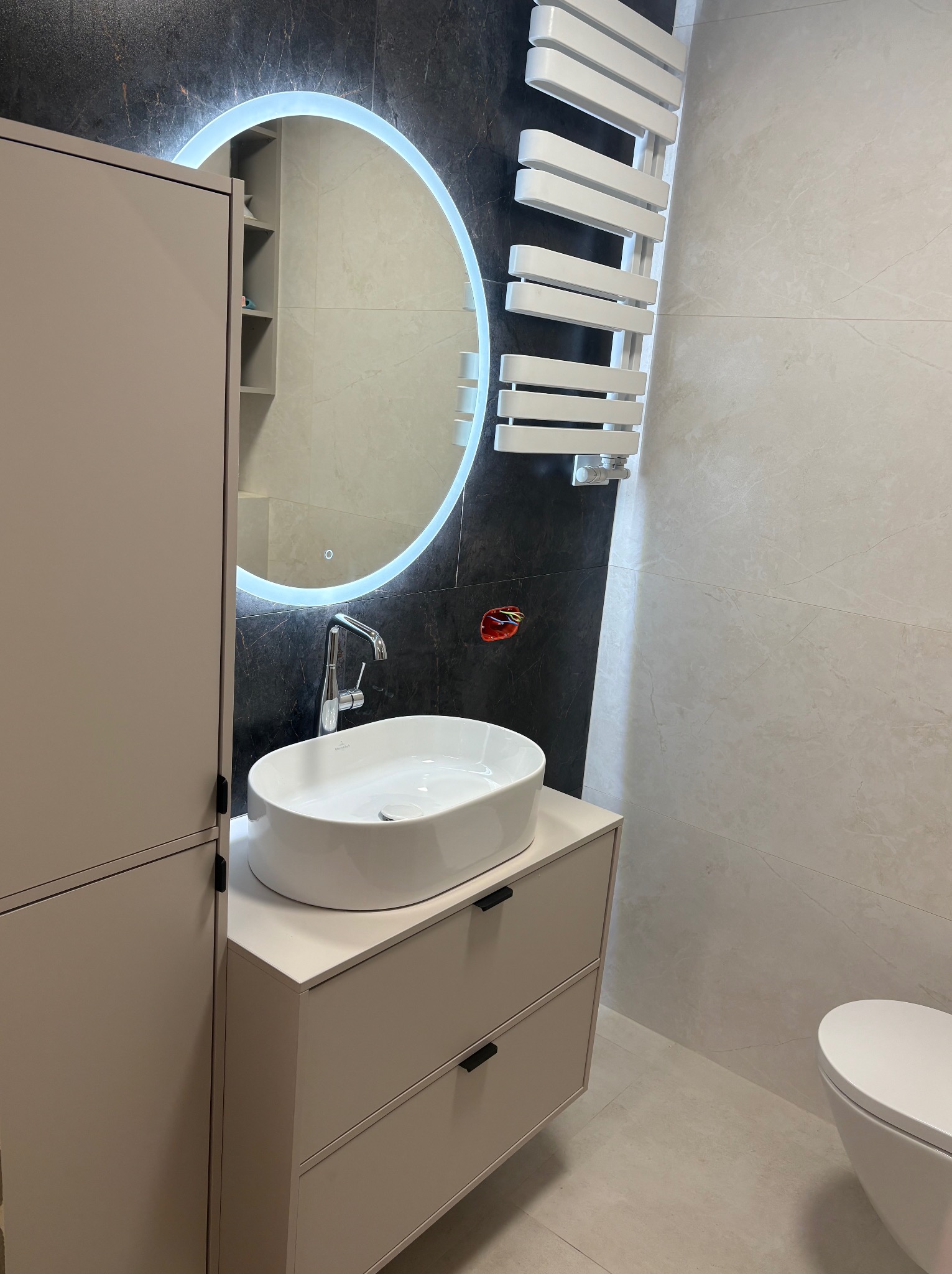 Nowoczesna łazienka z okrągłym lustrem LED, umywalką nablatową i grzejnikiem drabinkowym. Minimalistyczny design, beżowe szafki i płytki imitujące marmur.