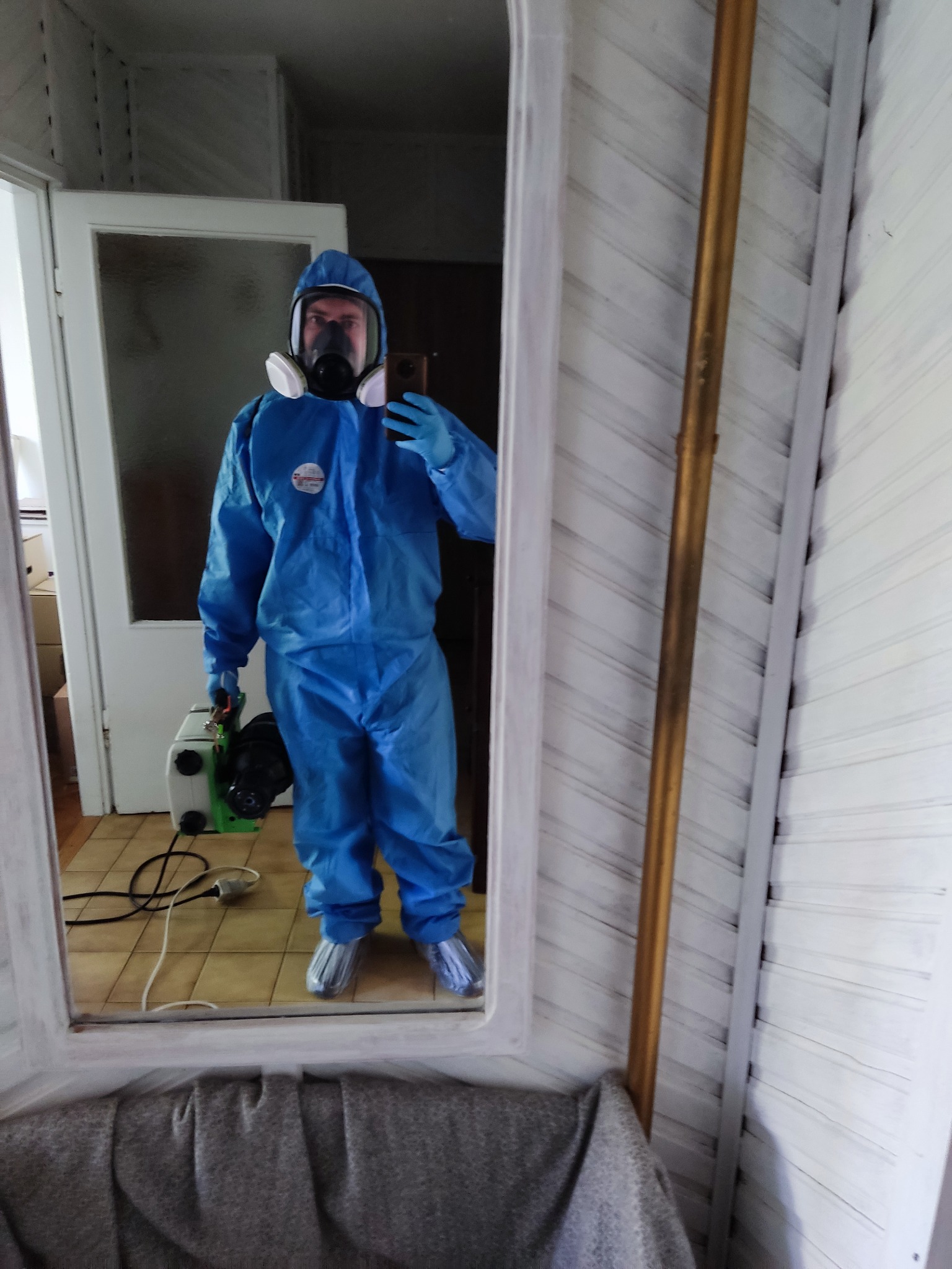 Pracownik w kombinezonie ochronnym, masce i rękawicach, trzymający urządzenie do zamgławiania, odbity w lustrze w pomieszczeniu z boazerią.