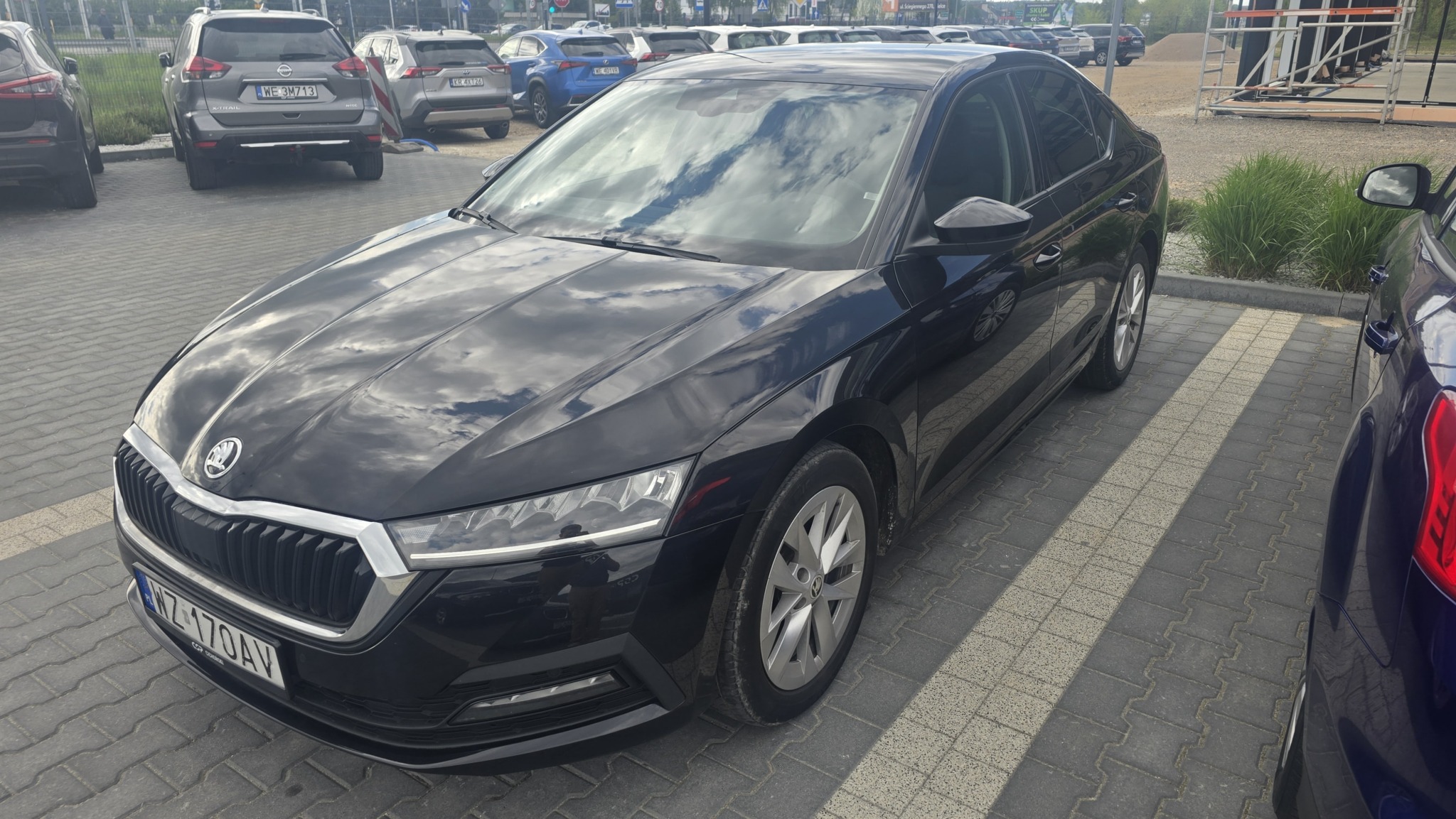 Elegancka, czarna Skoda Octavia zaparkowana na parkingu. Na masce odbijają się chmury. Widoczna tablica rejestracyjna i fragmenty innych aut w tle.
