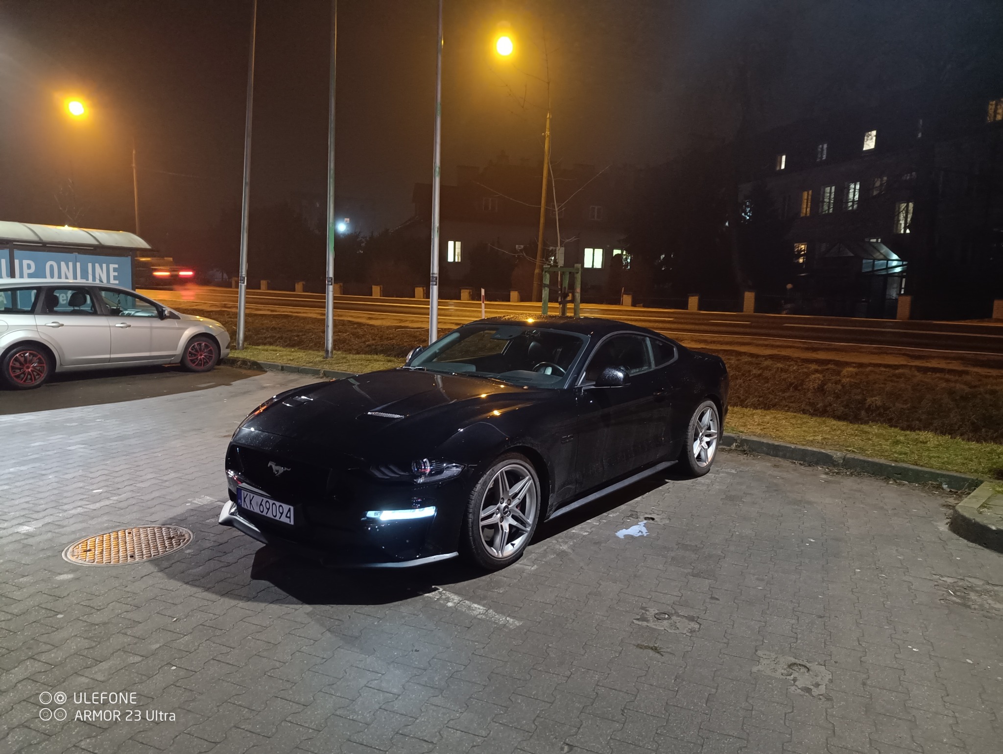 Czarny Ford Mustang, wynajem aut sportowych w Elblągu. Auto zaparkowane nocą na kostce brukowej, widoczne światła uliczne i budynek w tle. Tablica rejestracyjna widoczna.