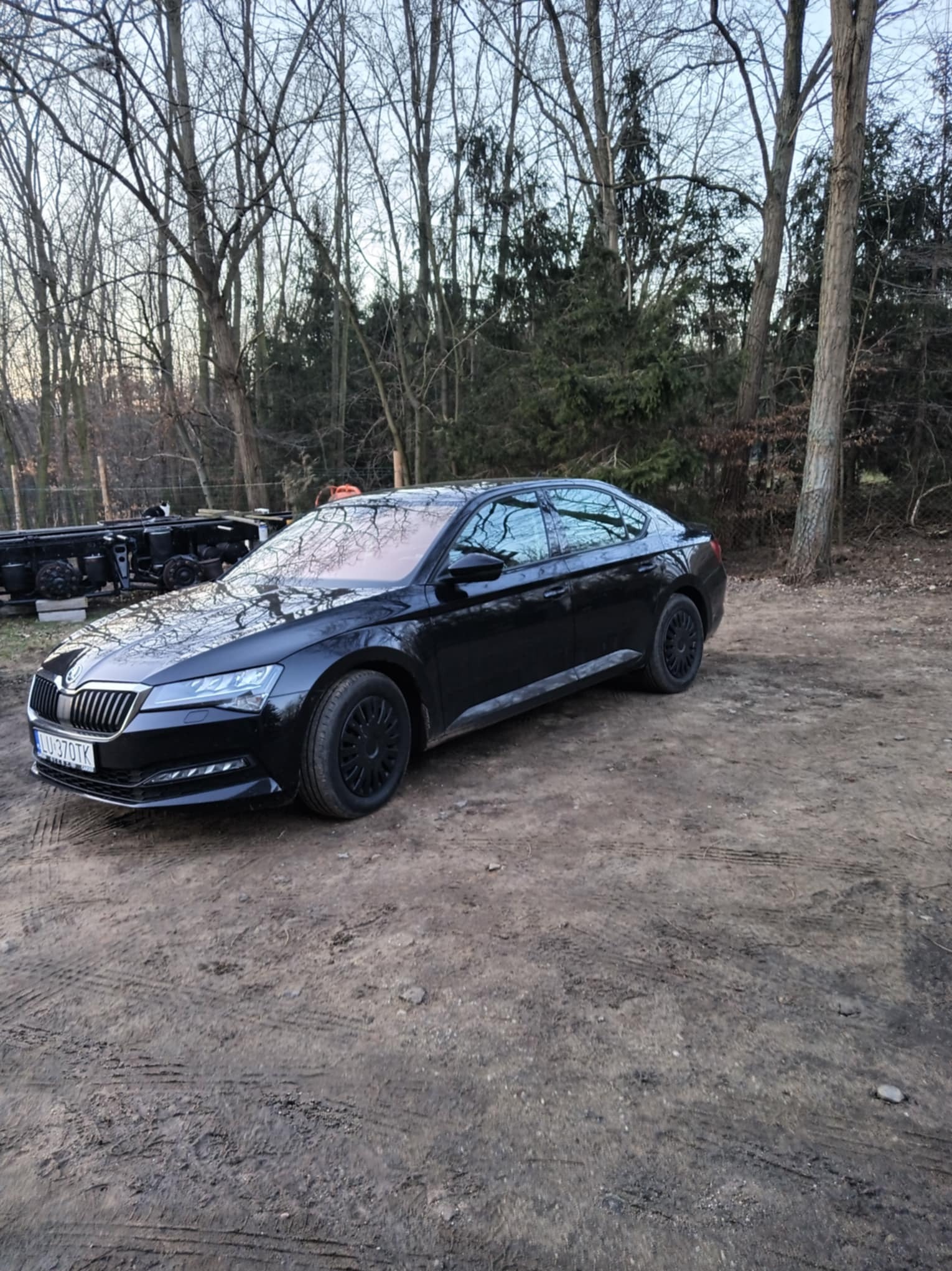 Czarna Skoda Superb z czarnymi felgami parkuje na ziemistej powierzchni, w tle drzewa i przyczepa. Auto z wypożyczalni.