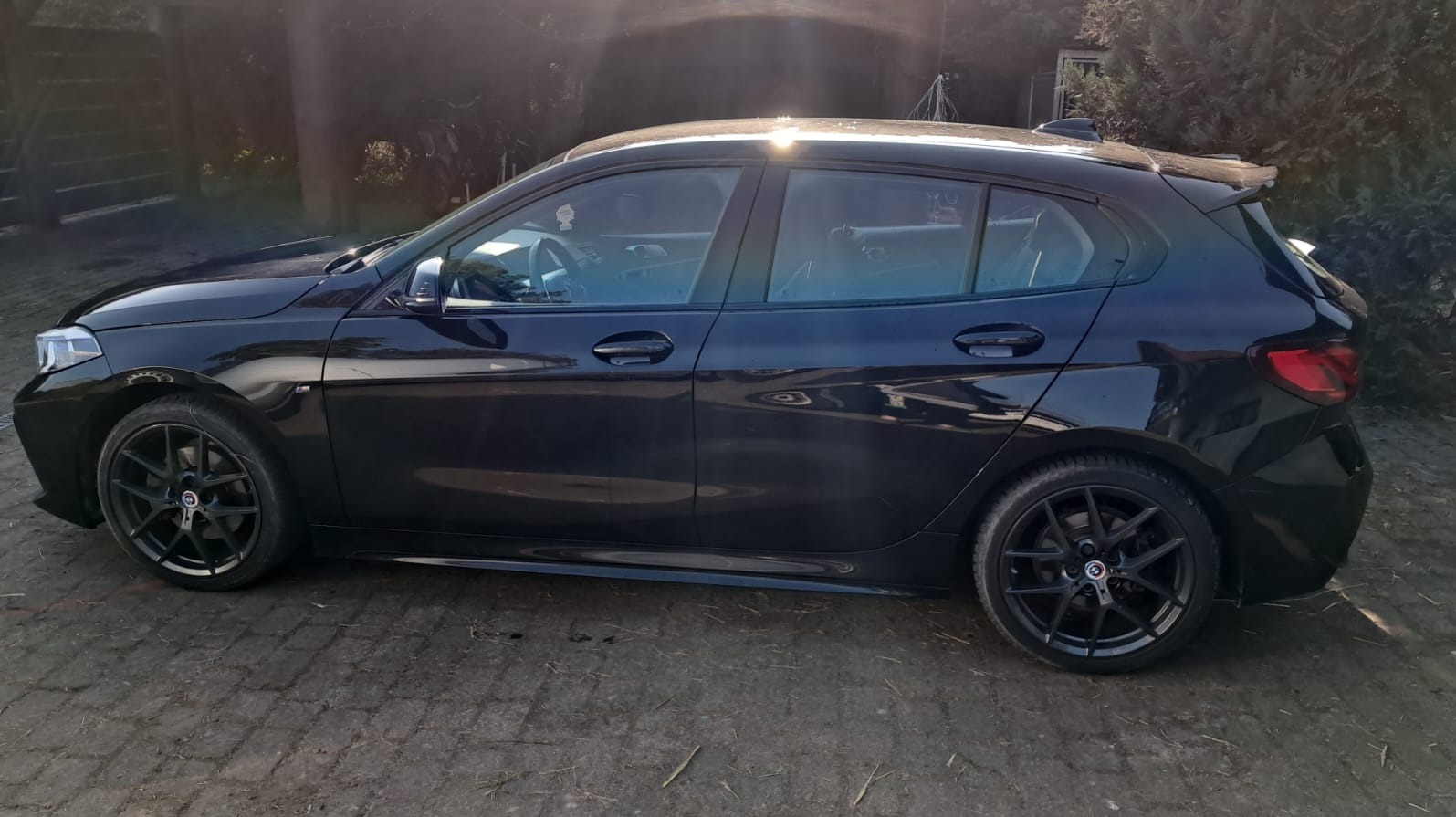 Czarny, zadbany hatchback BMW serii 1 z ciemnymi felgami, zaparkowany na brukowanym podjeździe w słoneczny dzień. Widok z boku, zarys drzew w tle.