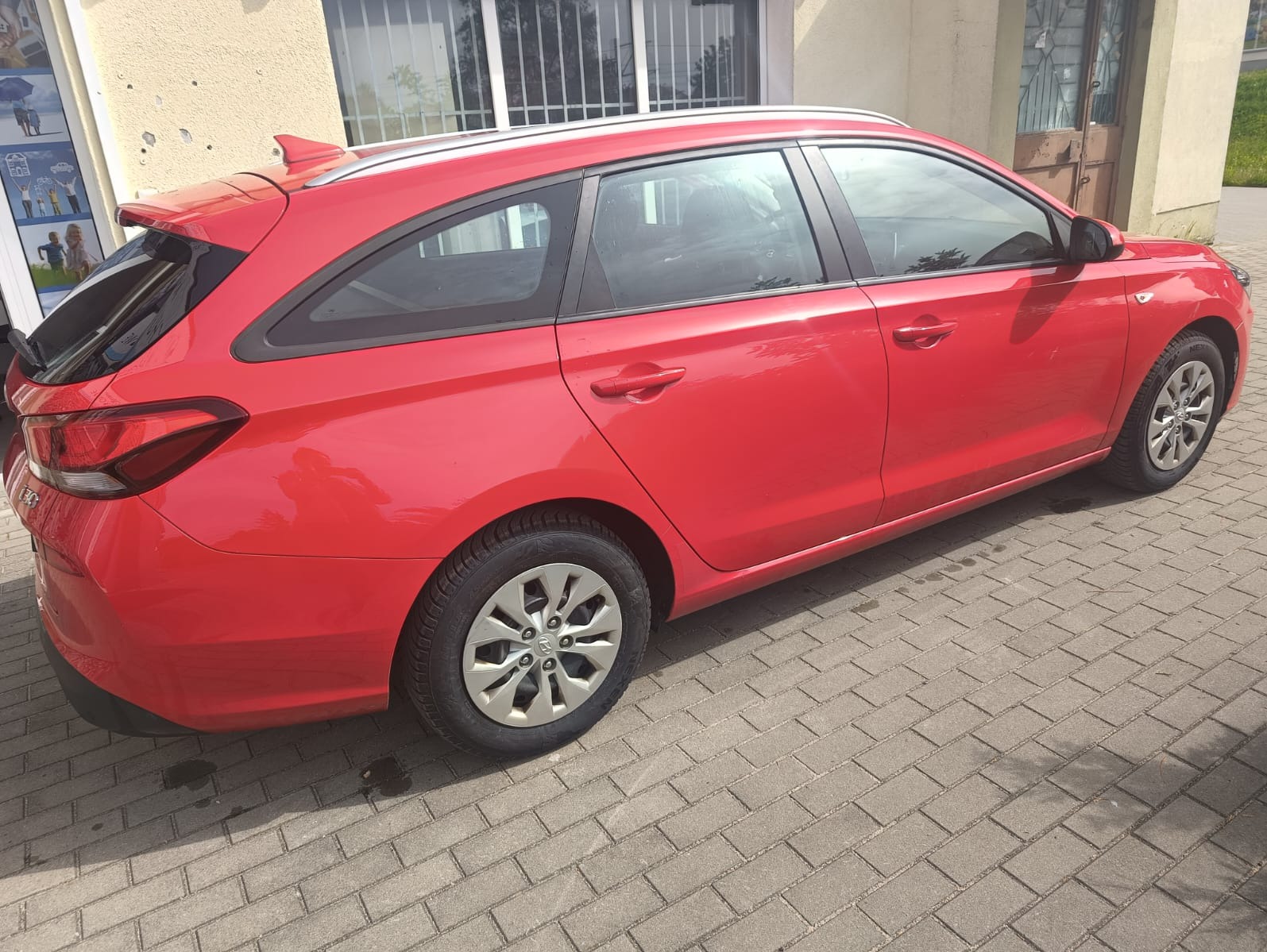 Czerwony samochód typu kombi, z relingami dachowymi, zaparkowany na brukowanej nawierzchni, widok z boku. Model z widocznymi detalami karoserii i felg.