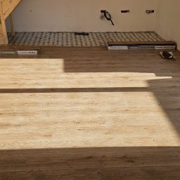 Pawel Wozniak usługi budowlano wykonczeniowe - Jasne panele podłogowe ułożone w pokoju z drewnianymi schodami i oknem. Część podłogi wyłożona podkładem pod panele. Widoczne paczki paneli Kronostep.