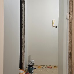 Pawel Wozniak usługi budowlano wykonczeniowe - Korytarz w trakcie malowania: świeżo pomalowane ściany na biało, widoczne puszki z farbą Dekoral Akrylit W, taśmy malarskie i narzędzia. Otwór drzwiowy w stanie surowym.