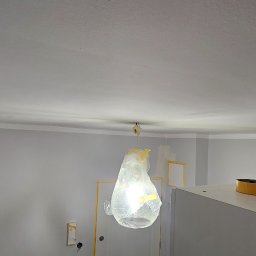 Pawel Wozniak usługi budowlano wykonczeniowe - Wnętrze po malowaniu: szare ściany, biały sufit z listwą, zabezpieczona lampa folią. Widoczne taśmy malarskie i otwór w ścianie. Ujęcie z dołu.
