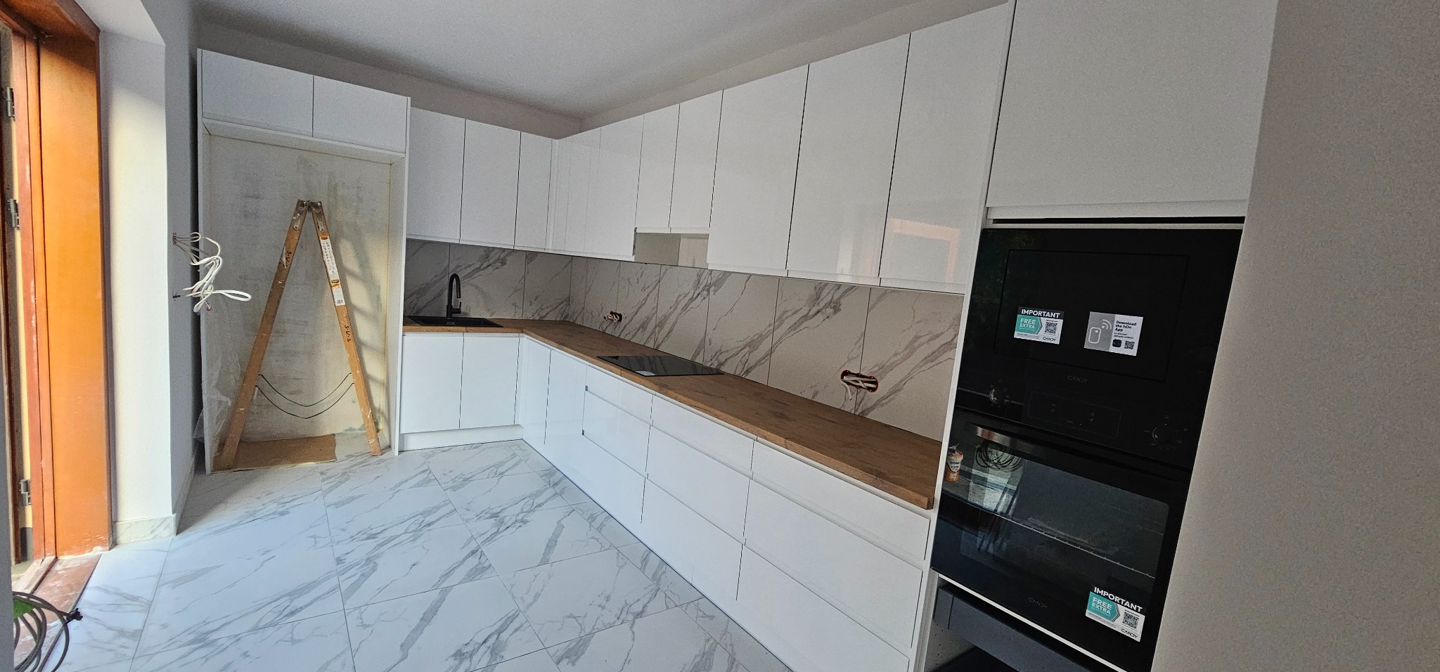 Nowoczesna kuchnia z białymi, lakierowanymi szafkami, drewnianym blatem i marmurowym backsplashiem. Widoczna drabina malarska sugeruje trwające prace wykończeniowe.