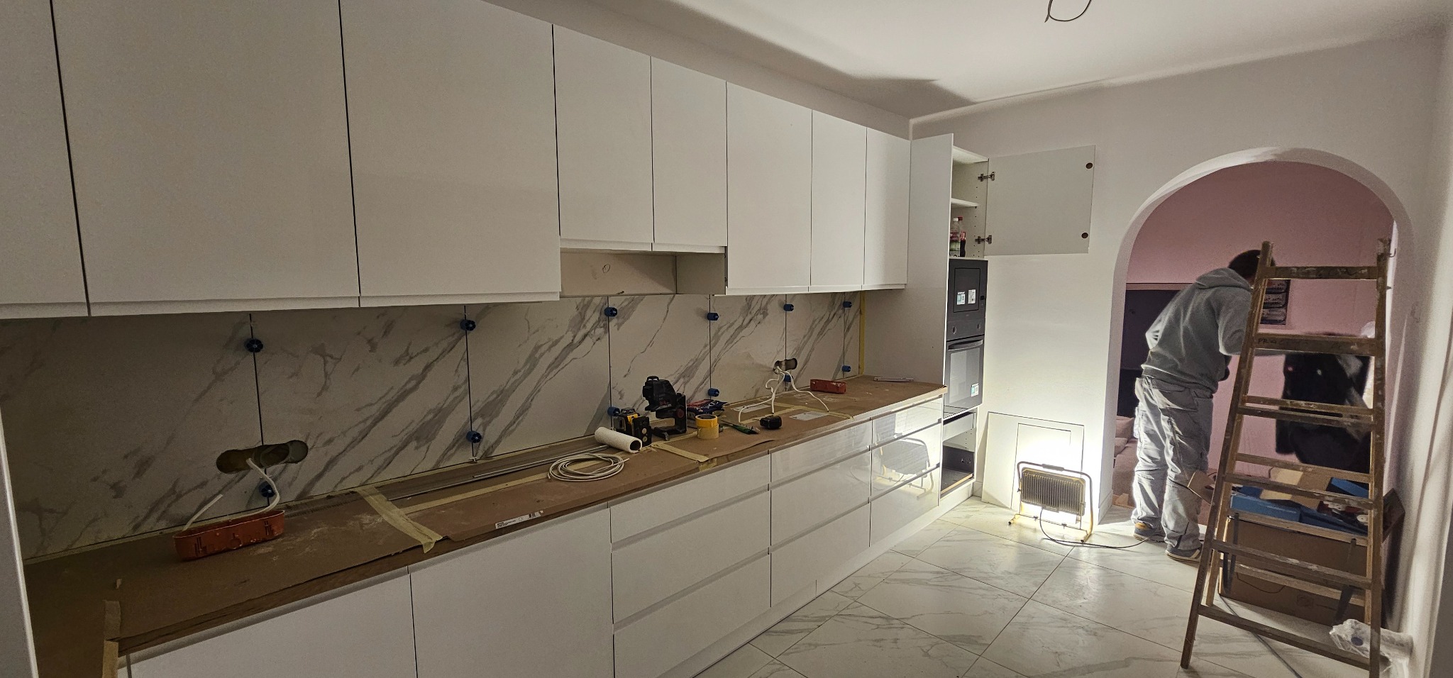 Biała kuchnia w trakcie montażu: widoczne płytki imitujące marmur z niebieskimi krzyżykami, częściowo zamontowane szafki i osoba pracująca w tle.