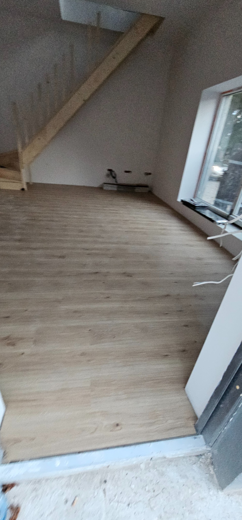 Jasne panele podłogowe w pokoju na poddaszu, widoczne fragmenty drewnianej konstrukcji dachu i okno. Świeżo położona podłoga w nowym wnętrzu.