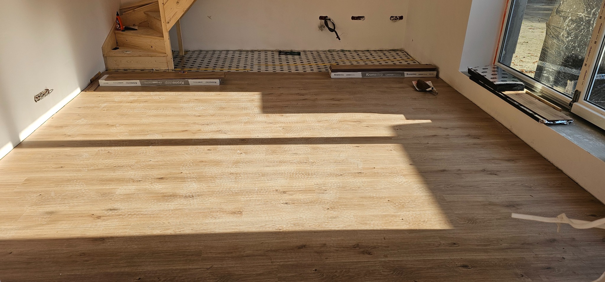 Jasne panele podłogowe ułożone w pokoju z drewnianymi schodami i oknem. Część podłogi wyłożona podkładem pod panele. Widoczne paczki paneli Kronostep.