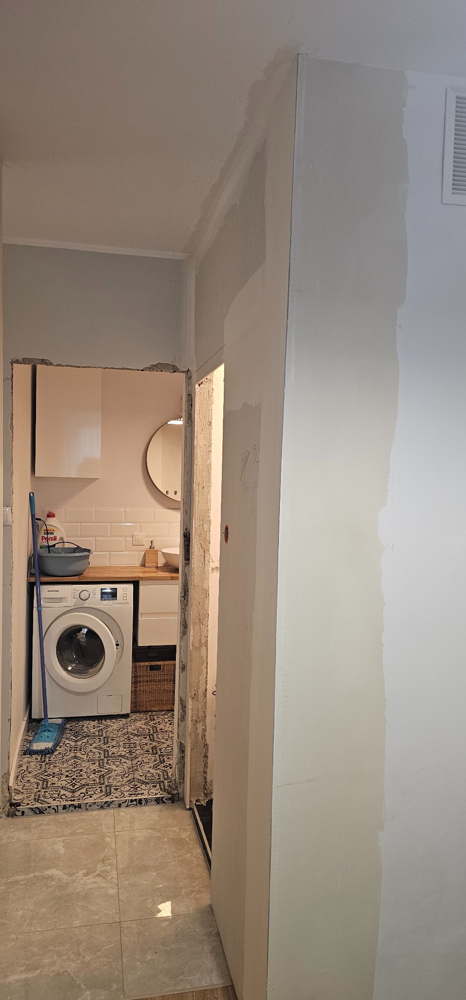 Wnętrze łazienki w trakcie remontu: widoczne fragmenty ściany po szpachlowaniu, pralka, umywalka i płytki. Ujęcie z perspektywy korytarza, z widocznym fragmentem wentylacji.