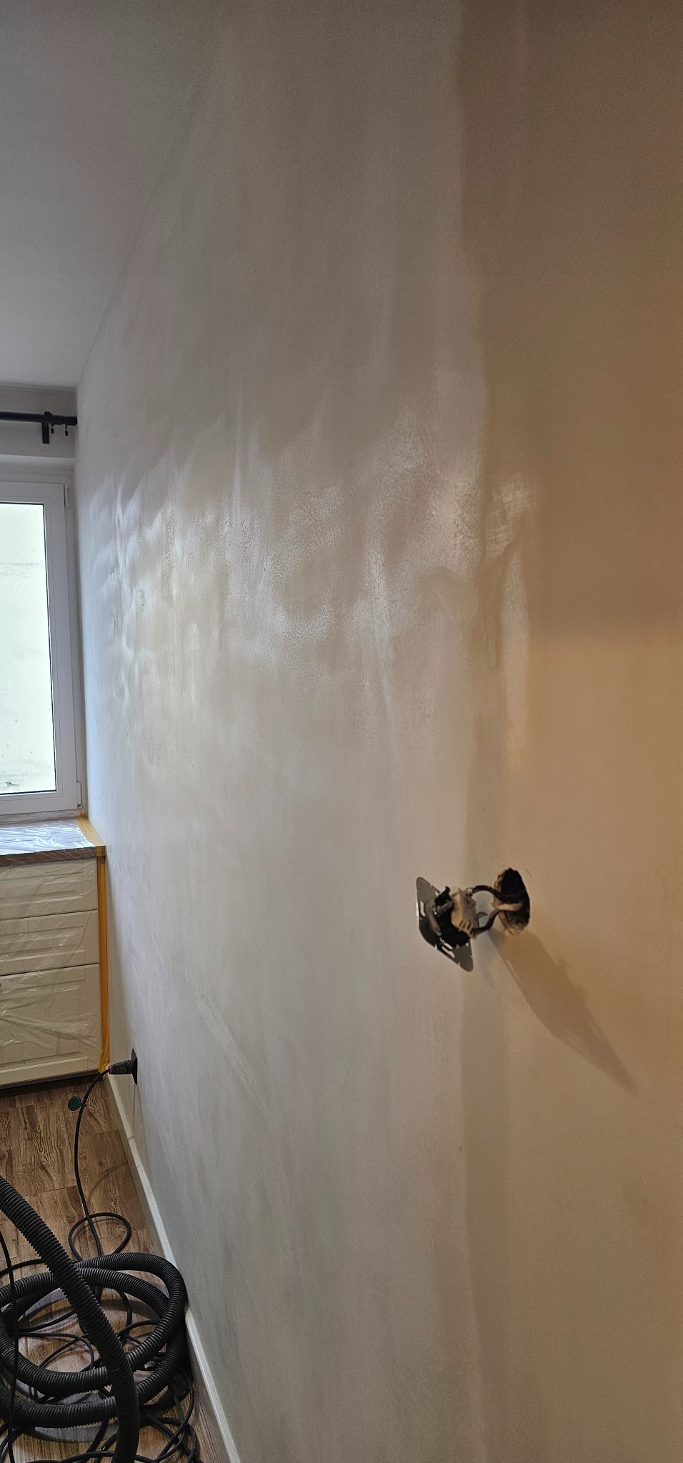 Ściana po szpachlowaniu z widocznym gniazdkiem elektrycznym, fragmentem okna i podłogi. Widoczne ślady po szpachlowaniu. W tle odkurzacz.