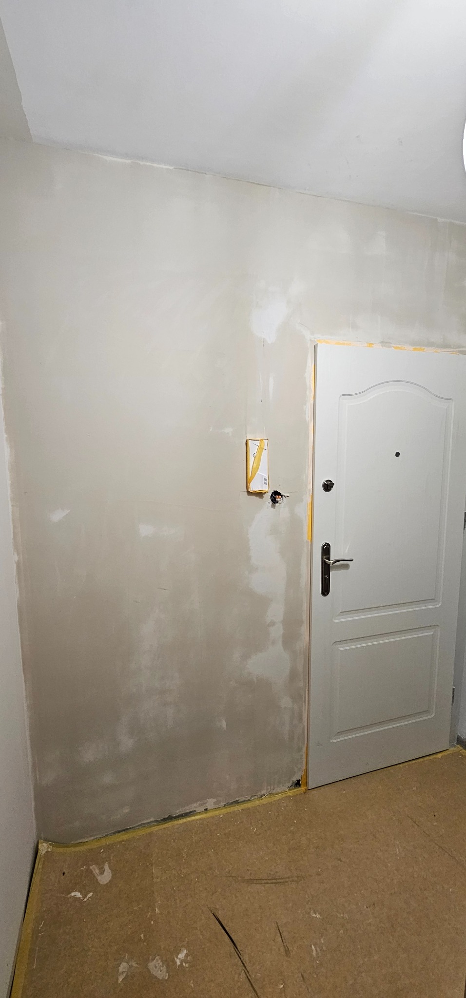 Ściana po szpachlowaniu, przygotowana do malowania. Widoczne ślady po łatach i taśma malarska przy futrynie drzwi wejściowych. Gniazdko elektryczne zabezpieczone.