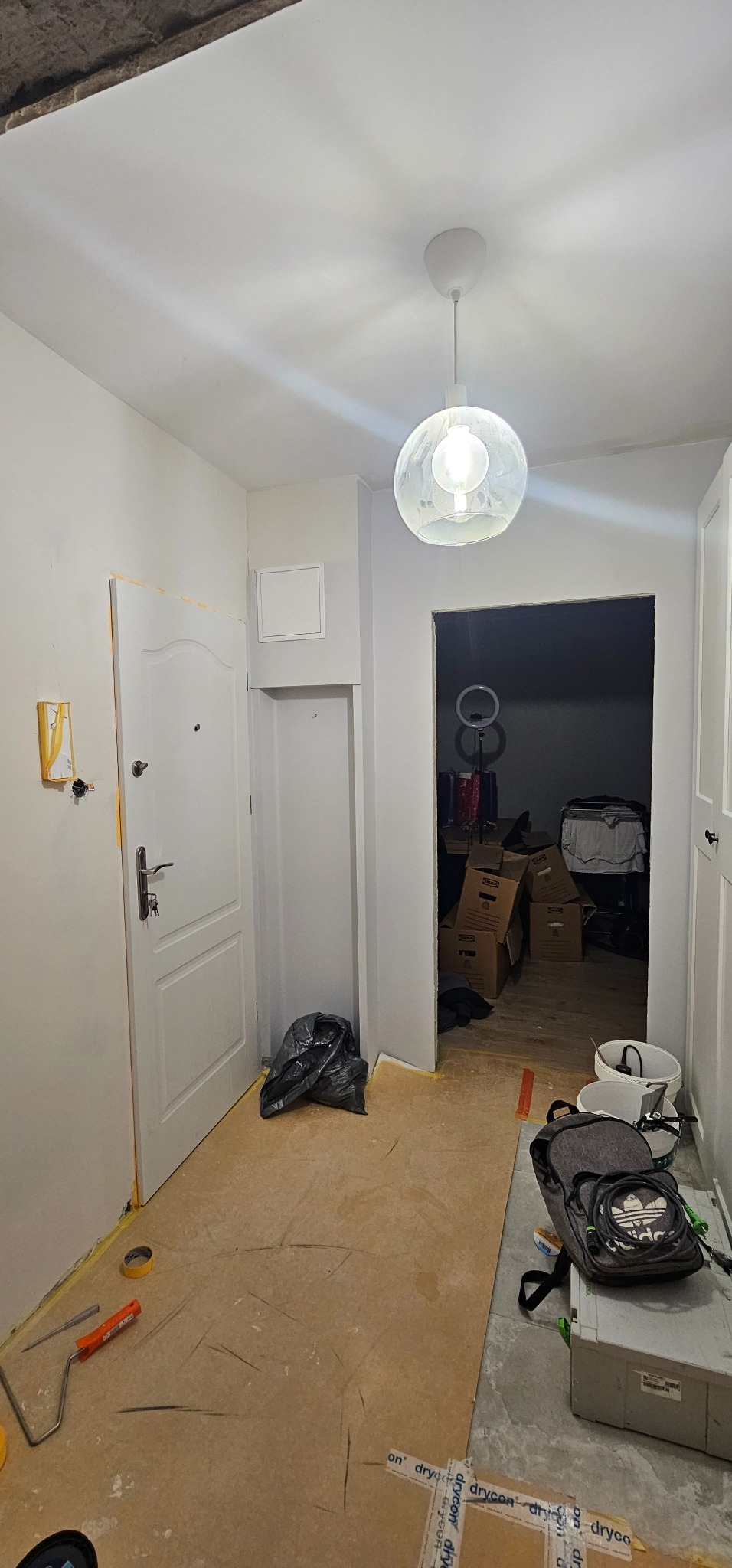 Korytarz w trakcie prac wykończeniowych: zabezpieczona podłoga, świeżo pomalowane ściany, widoczne narzędzia i akcesoria malarskie. W tle otwarte drzwi do innego pomieszczenia.