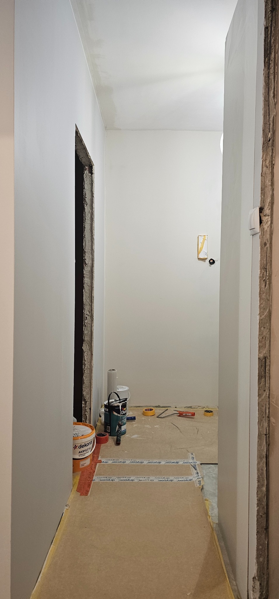 Korytarz w trakcie malowania: świeżo pomalowane ściany na biało, widoczne puszki z farbą Dekoral Akrylit W, taśmy malarskie i narzędzia. Otwór drzwiowy w stanie surowym.