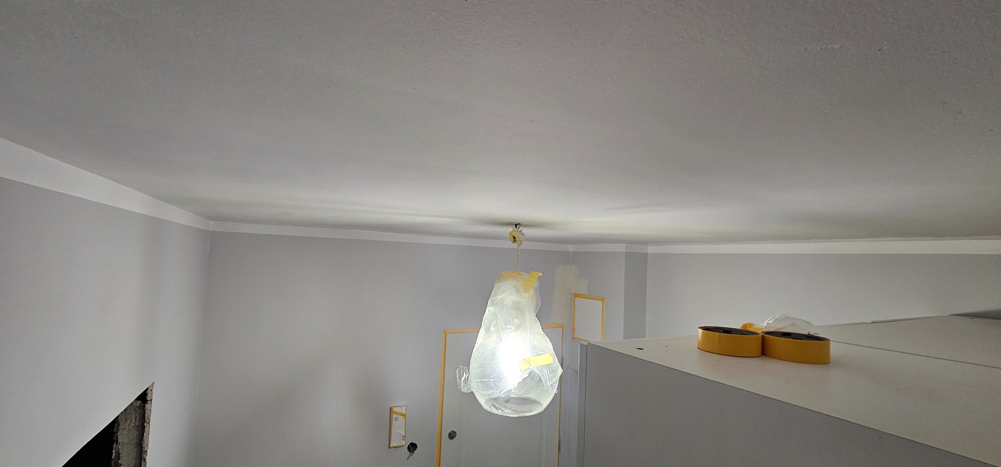 Wnętrze po malowaniu: szare ściany, biały sufit z listwą, zabezpieczona lampa folią. Widoczne taśmy malarskie i otwór w ścianie. Ujęcie z dołu.
