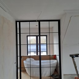 Remtech Michał Bryndal - Nowoczesna, czarna, metalowa konstrukcja z szybami, prawdopodobnie drzwi lub ścianka działowa, w surowym wnętrzu. Widoczna drabina i zabezpieczona folią wanna.