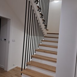 Remtech Michał Bryndal - Minimalistyczne schody drewniane z czarną, pionową balustradą w nowoczesnym wnętrzu. Jasne stopnie i białe ściany, oświetlenie sufitowe.