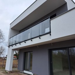 Remtech Michał Bryndal - Nowoczesny balkon z przeszkloną balustradą w minimalistycznym budynku, widok z zewnątrz. Szklane panele osadzone w czarnych profilach, tło stanowią drzewa bez liści.