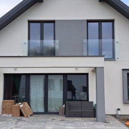 Remtech Michał Bryndal - Nowoczesny dom z balkonem zabezpieczonym szklaną balustradą. Widoczne duże okna, szare elementy elewacji i kostka brukowa. W oknie siedzi pies.