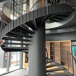 Remtech Michał Bryndal - Szare metalowe schody spiralne z centralnym słupem, widoczne z dołu, w surowym wnętrzu z zabezpieczeniami podłogi, podkreślające minimalistyczny design.