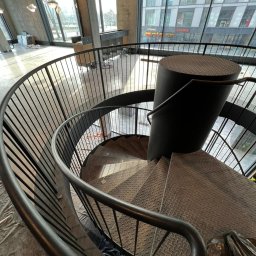 Remtech Michał Bryndal - Spiralne schody metalowe z czarną balustradą, widok z góry na ażurową konstrukcję. W tle duże okna z widokiem na budynek miejski, widać elementy wykończeniowe wnętrza.