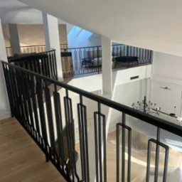 Czarna, metalowa balustrada schodowa z pionowymi, prostokątnymi elementami dekoracyjnymi, widok z góry, jasne wnętrze, drewniana podłoga, elementy zabezpieczone folią.