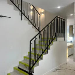 Nowoczesne schody z czarną, metalową balustradą o unikalnym wzorze. Stopnie zabezpieczone taśmą malarską, marmurowa podłoga w tle. Widok na jasne wnętrze domu.