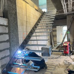 Remtech Michał Bryndal - Spawanie metalowych schodów w surowym wnętrzu budynku. Widoczny spawacz w trakcie pracy, iskry, elementy konstrukcyjne schodów i sprzęt spawalniczy.