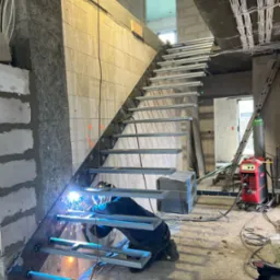Spawanie metalowych schodów w surowym wnętrzu budynku. Widoczny spawacz w trakcie pracy, iskry, elementy konstrukcyjne schodów i sprzęt spawalniczy.