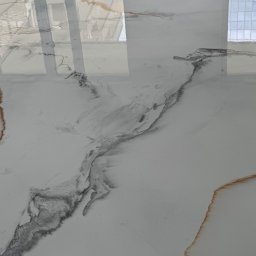 RawEpoxyStudio - Błyszcząca, biała posadzka żywiczna z szarymi i złotymi żyłkami, przypominająca marmur. Widoczne odbicie okna w gładkiej powierzchni.