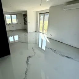 Biała, błyszcząca posadzka żywiczna z szarymi wzorami marmuru w nowym, jasnym pomieszczeniu z oknami i klimatyzacją. Widoczne niedociągnięcia wykończeniowe ścian.