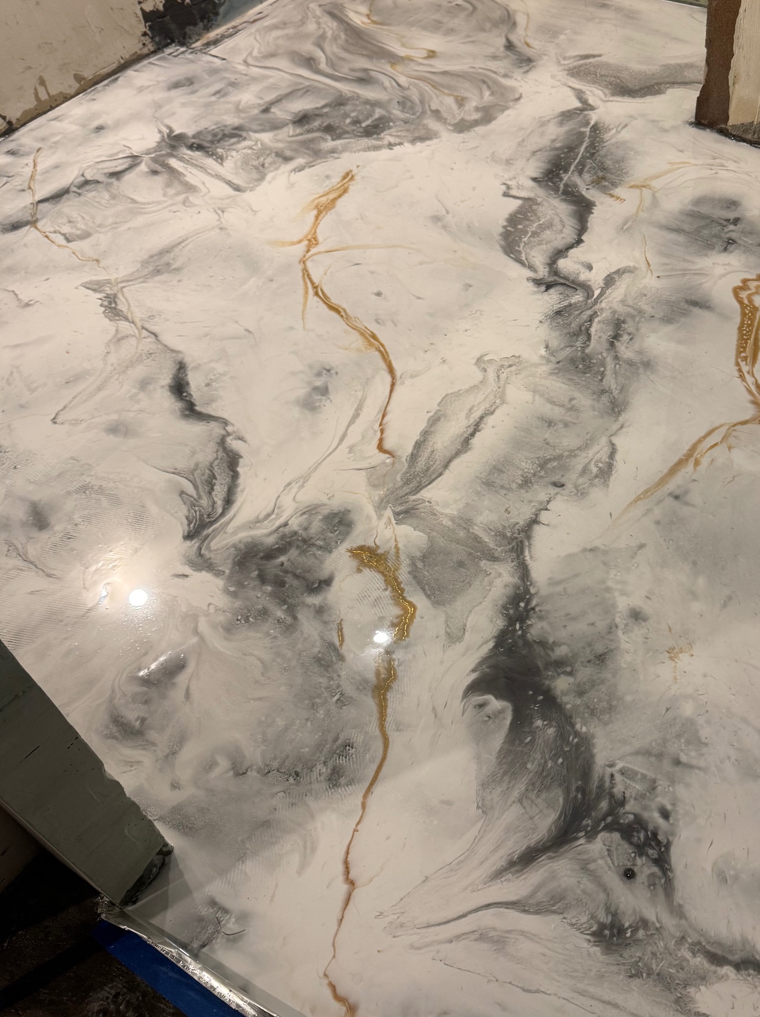 Błyszcząca posadzka żywiczna w odcieniach bieli, szarości i złota, imitująca marmur, świeżo wylana, z widocznymi detalami wykończenia przy ścianie.