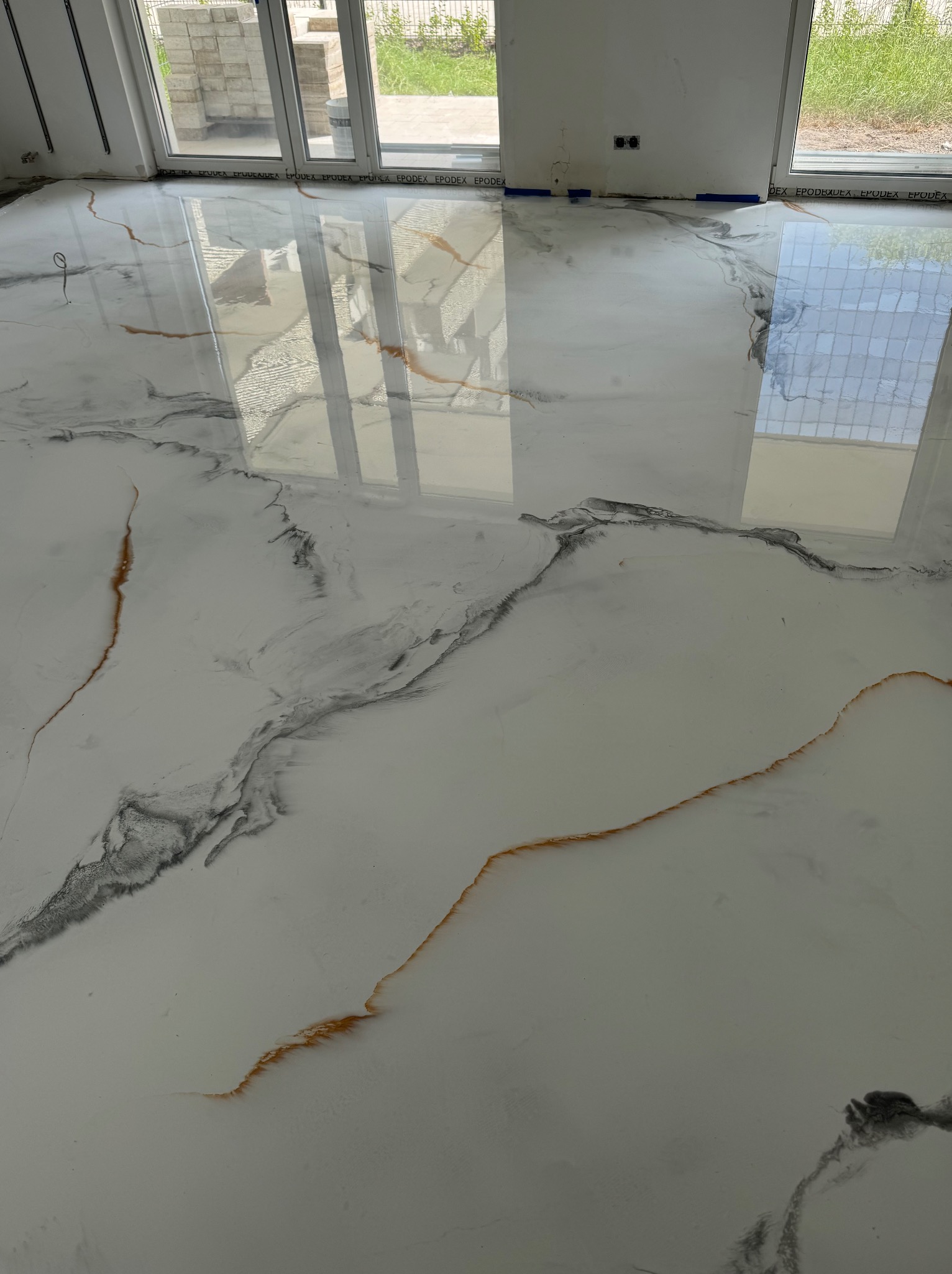 Błyszcząca, biała posadzka z żywicy epoksydowej w nowym wnętrzu, z szarymi i złotymi żyłkami imitującymi marmur, odbijająca światło z dużych okien.