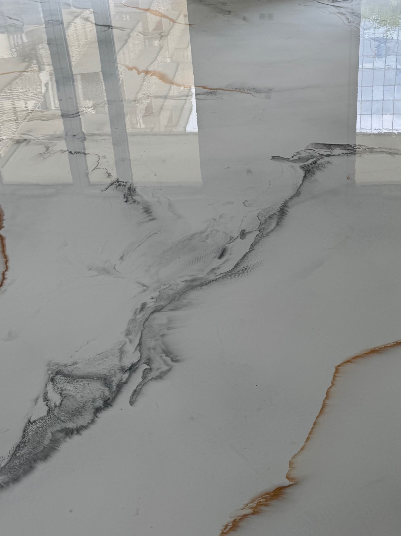 Błyszcząca, biała posadzka żywiczna z szarymi i złotymi żyłkami, przypominająca marmur. Widoczne odbicie okna w gładkiej powierzchni.