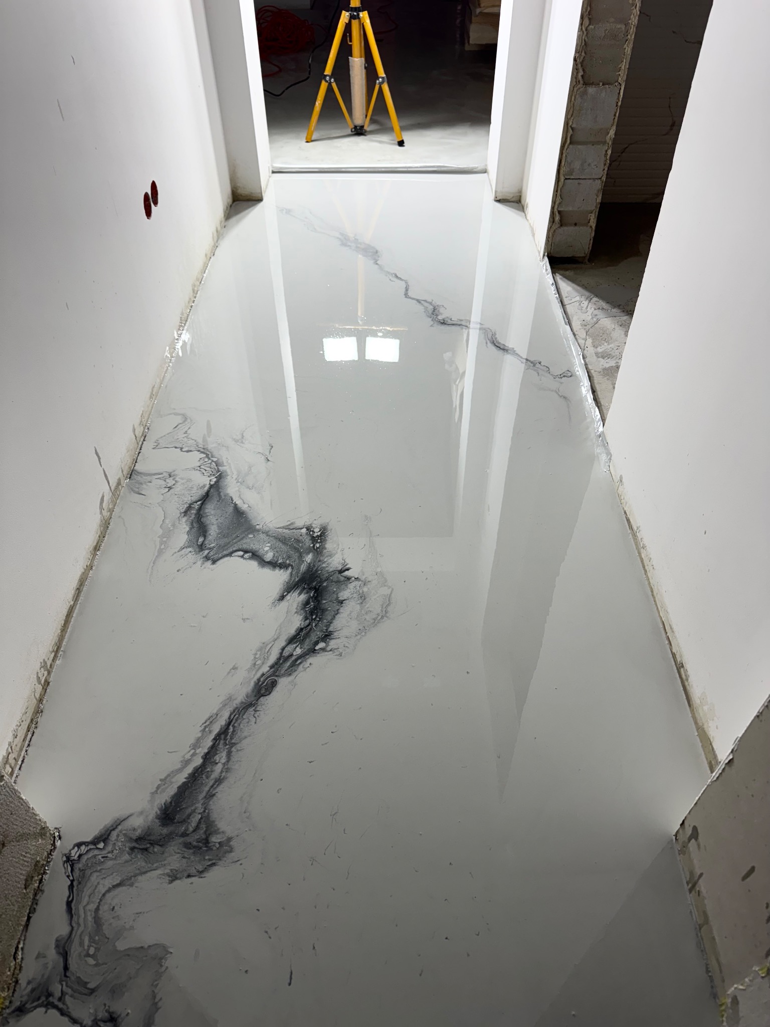 Błyszcząca, jasnoszara posadzka z efektem marmuru w korytarzu. Widoczny trójnóg oświetleniowy w tle i surowe wykończenie ścian.