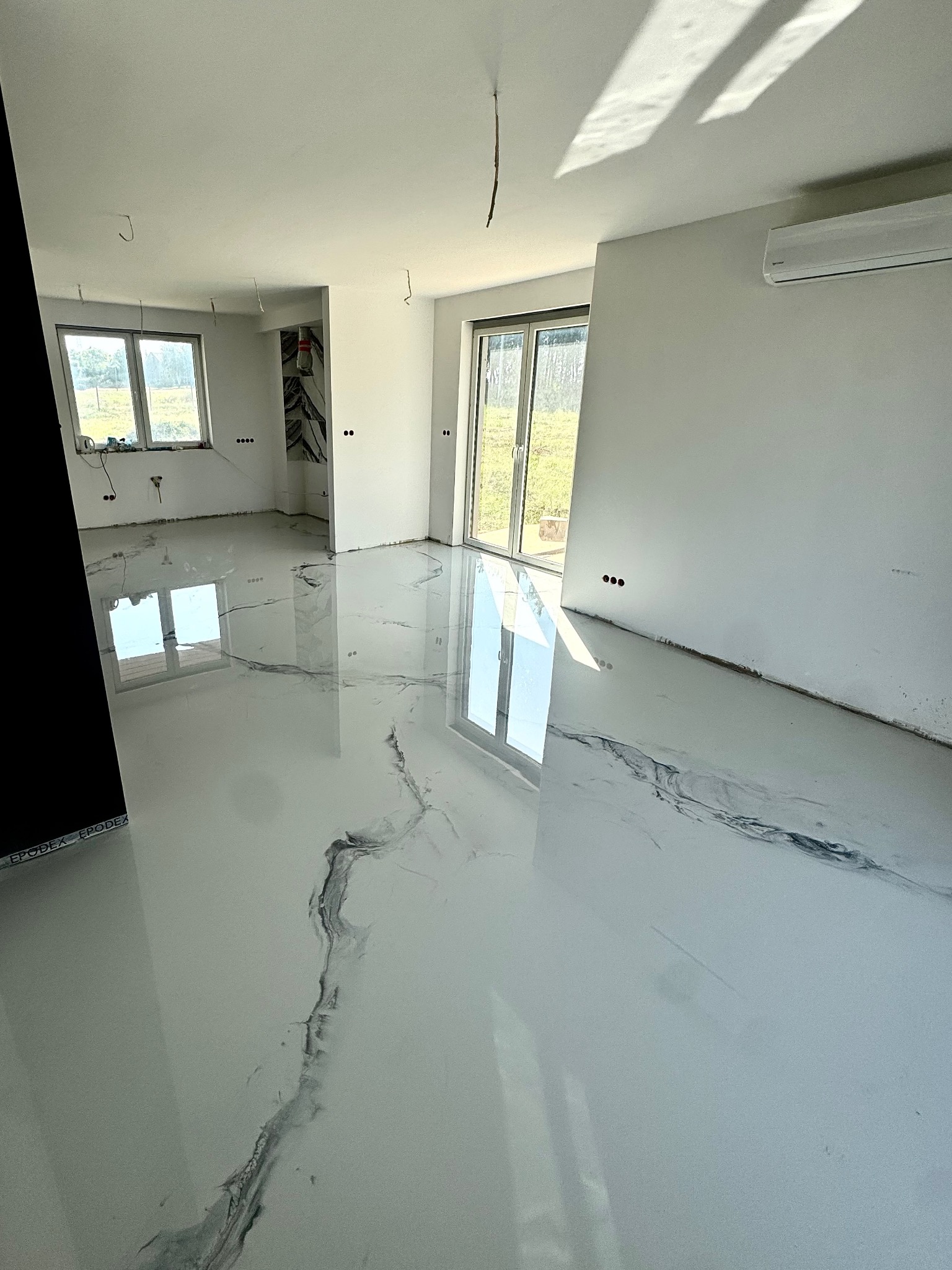 Biała, błyszcząca posadzka żywiczna z szarymi wzorami marmuru w nowym, jasnym pomieszczeniu z oknami i klimatyzacją. Widoczne niedociągnięcia wykończeniowe ścian.