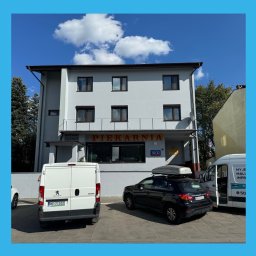 KORWEK Michał Korwek - Dwupiętrowy budynek piekarni z szarym tynkiem, brązowymi ramami okiennymi i żółtym napisem. Na parkingu zaparkowane samochody dostawcze. Błękitne niebo w tle.