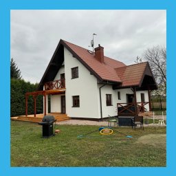 KORWEK Michał Korwek - Biały dom z czerwoną dachówką i drewnianymi elementami wykończeniowymi, widoczny z perspektywy ogrodu. Dom po renowacji, z nową elewacją i tarasem.