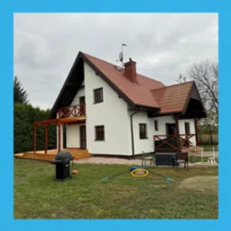 Biały dom z czerwoną dachówką i drewnianymi elementami wykończeniowymi, widoczny z perspektywy ogrodu. Dom po renowacji, z nową elewacją i tarasem.