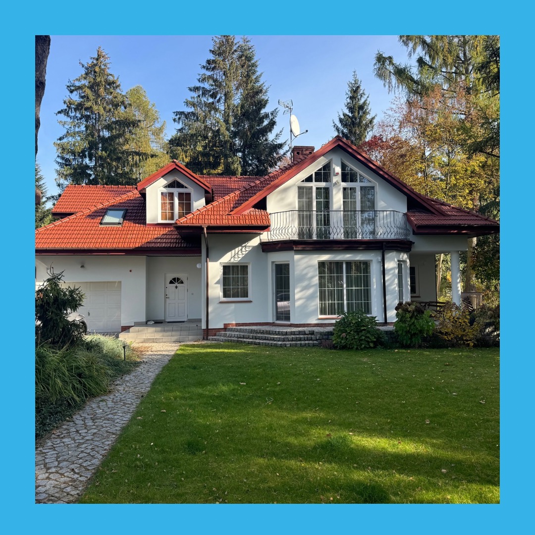 Biały dom jednorodzinny z czerwoną dachówką, balkonem i garażem, otoczony zielenią. Widoczny kamienny chodnik i drzewa w tle. Słoneczny dzień.
